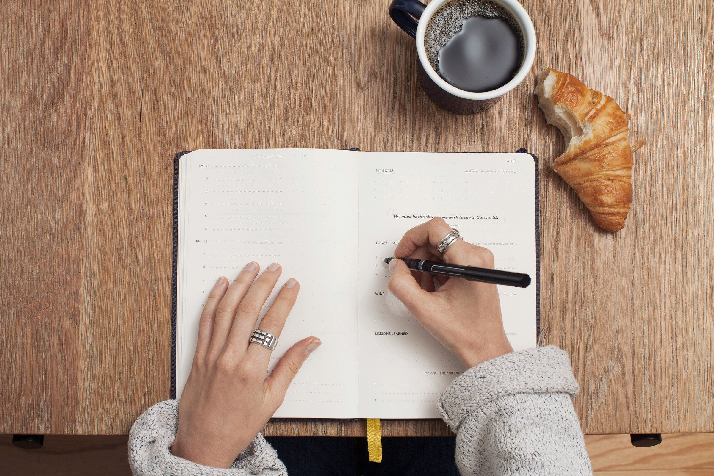 Administrative professional writing to do list in a planner while sipping on coffee and a croissant. 